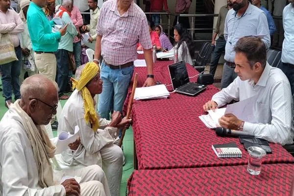 मेज के सामने बैठे ग्रामीणों से आवेदन लेते अधिकारी और आसपास खड़े लोग